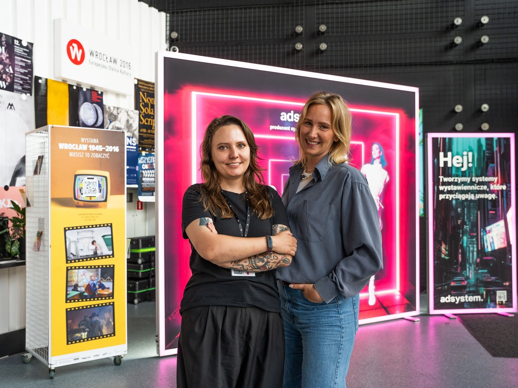 Zdjęcie przedstawia dwie kobiety stojące przed podświetlaną ścianką reklamową adsystem podczas wydarzenia we Wrocławiu.