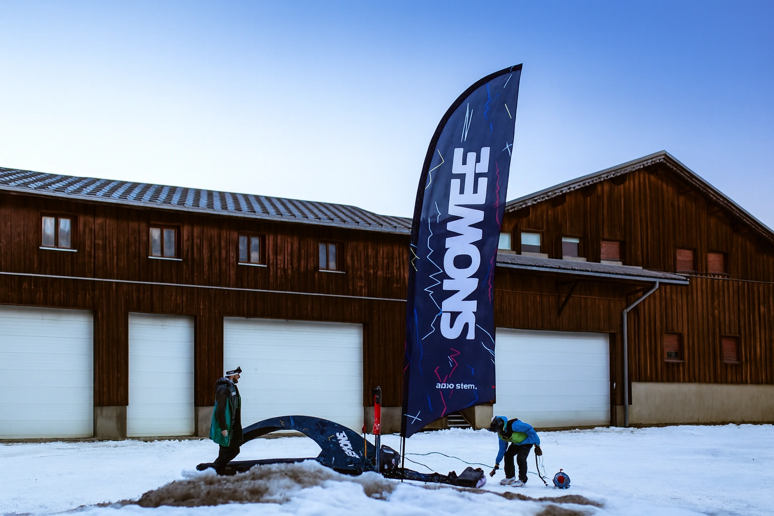 Flaga reklamowa SNOWEE z logo adsystem ustawiona na śniegu przed drewnianym budynkiem podczas przygotowań do zimowego eventu.