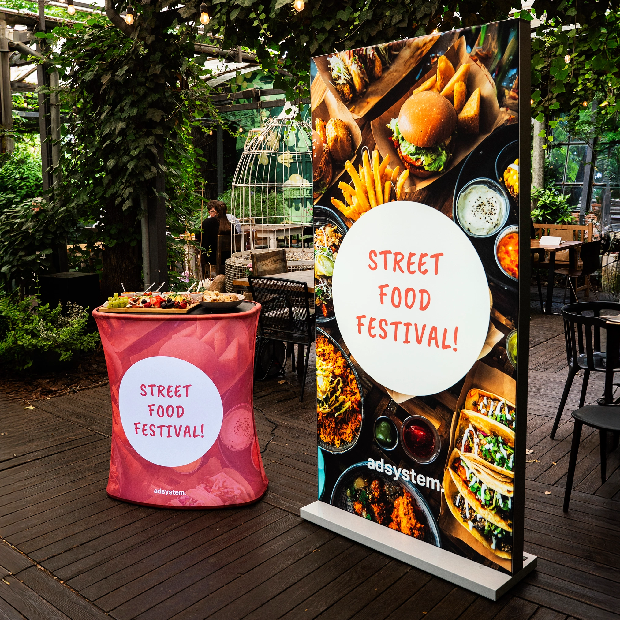 Trybunka i ścianka reklamowa z nadrukiem „Street Food Festival!” ustawione obok siebie w ogrodowej strefie gastronomicznej podczas plenerowego wydarzenia.