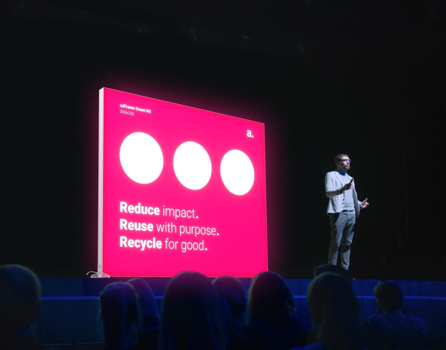 file-meta-description.apresentadordepéaoladodeumpainelpublicitárioiluminadocom-ledad-frame-smart-r3emumpalcoescuro,destacandoamensagem"-reduce.-reuse.-recycle."diantedeumpúblico.