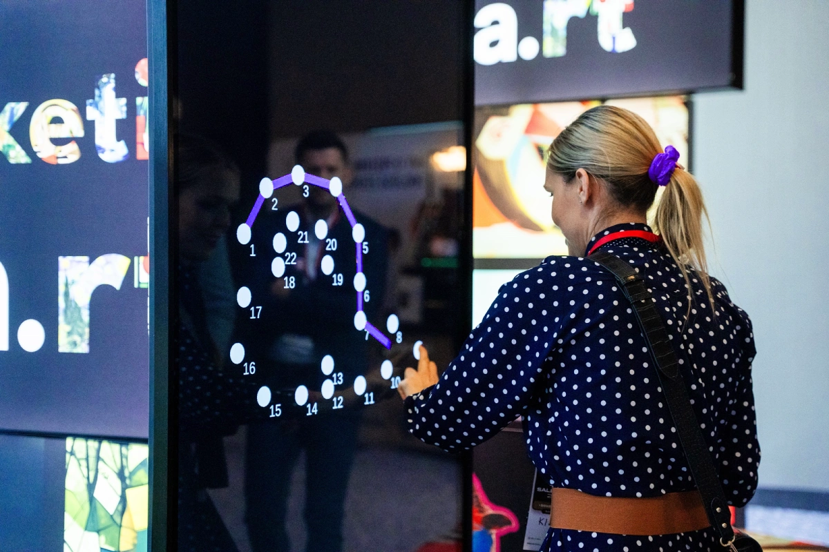 Uczestniczka eventu korzysta z interaktywnego ekranu dotykowego, wykonując zadanie typu „połącz kropki” w ramach angażującej grywalizacji targowej.