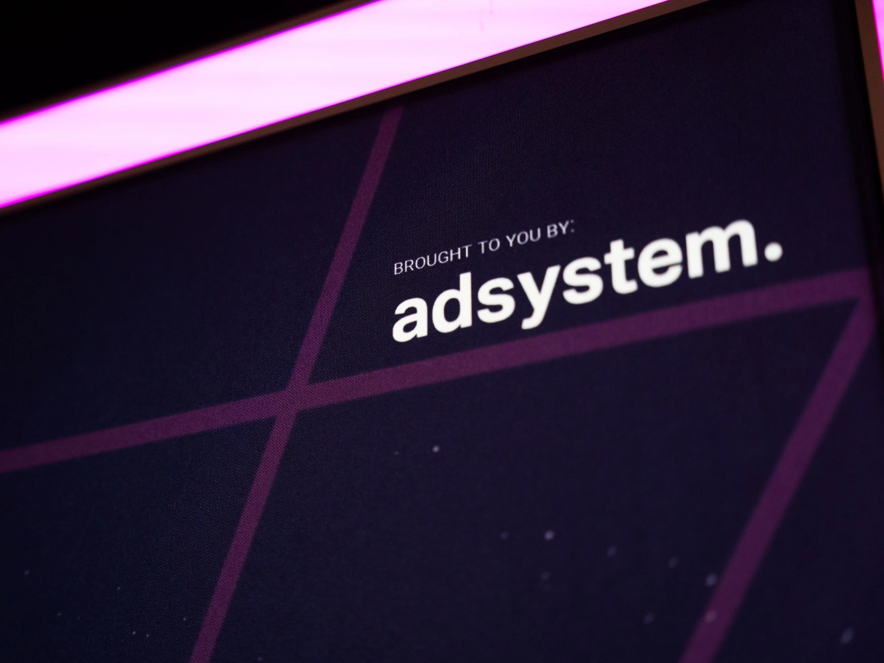 Zdjęcie przedstawia zbliżenie na fragment grafiki z logo adsystem i napisem „BROUGHT TO YOU BY: adsystem.” na tle podświetlanego kasetonu.