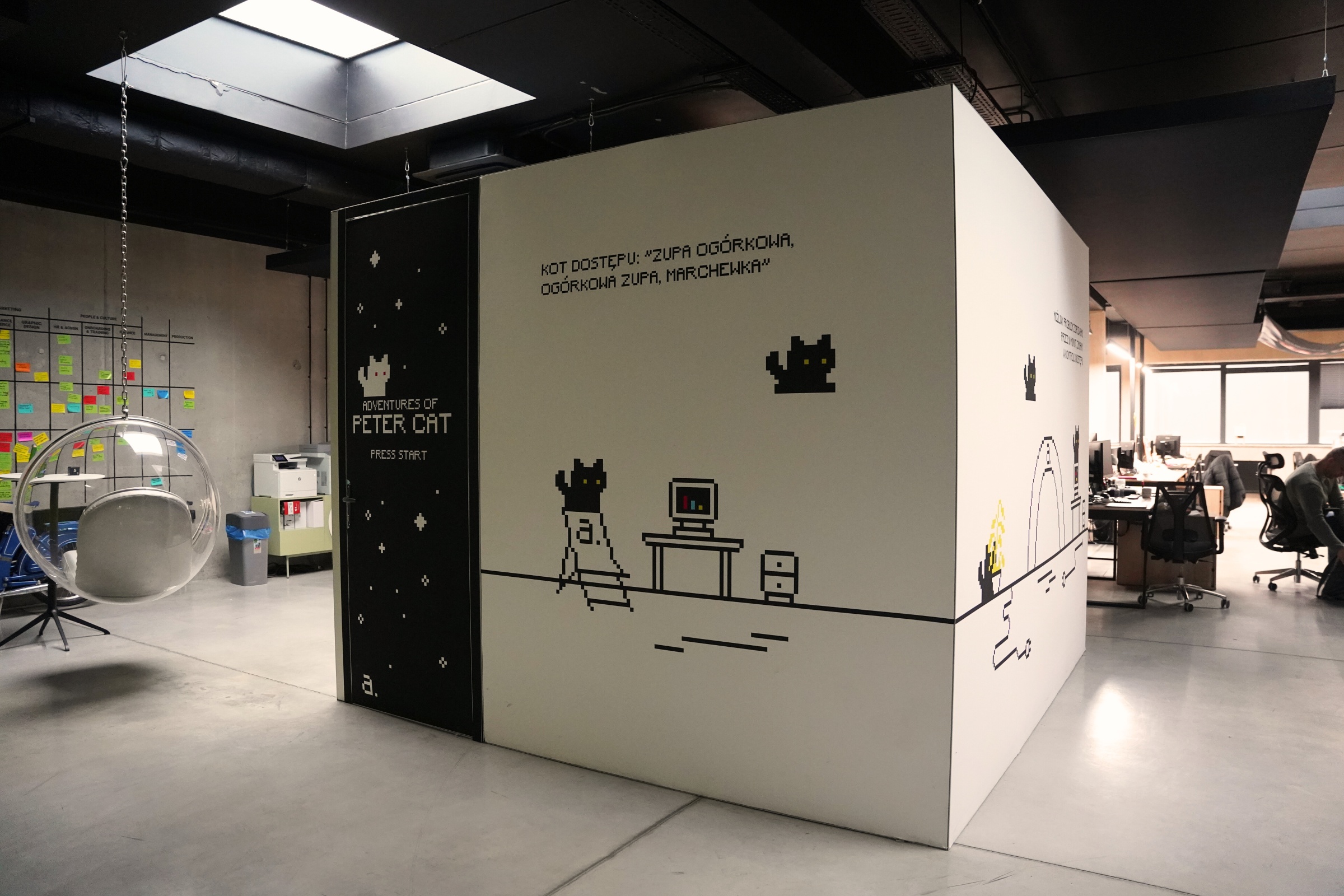 Nowoczesne biuro z zabudową ozdobioną grafiką w stylu pixel art, przedstawiającą kota i komputer; na ścianie napis „Adventures of Peter Cat”; obok designerski wiszący fotel i otwarta przestrzeń biurowa.