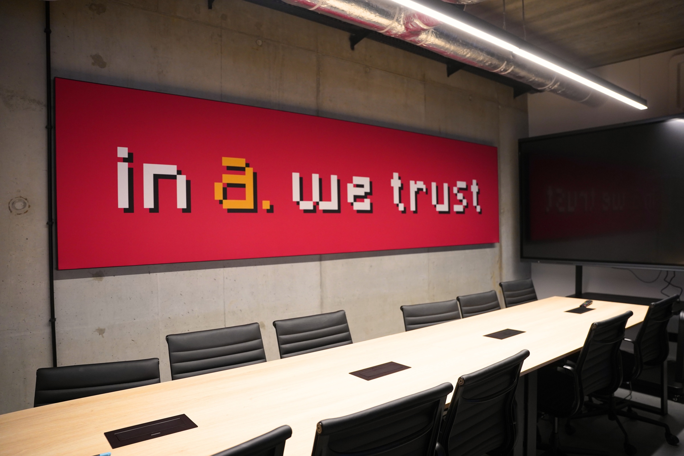 Sala konferencyjna z długim stołem i czarnymi krzesłami, na betonowej ścianie czerwona grafika z napisem „in a. we trust” w stylu pixel art, nad nią widoczne industrialne oświetlenie i instalacje sufitowe.