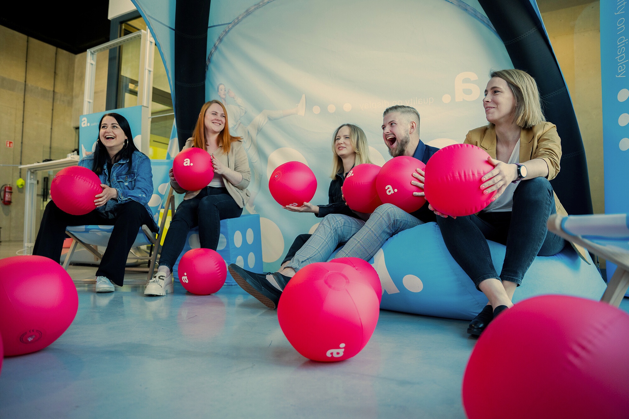 Grupa uśmiechniętych osób siedzi na pufach i leżakach w przestrzeni eventowej z logo „a.”, trzymając różowe piłki plażowe z nadrukiem; w tle niebieska ścianka reklamowa i dmuchana konstrukcja.