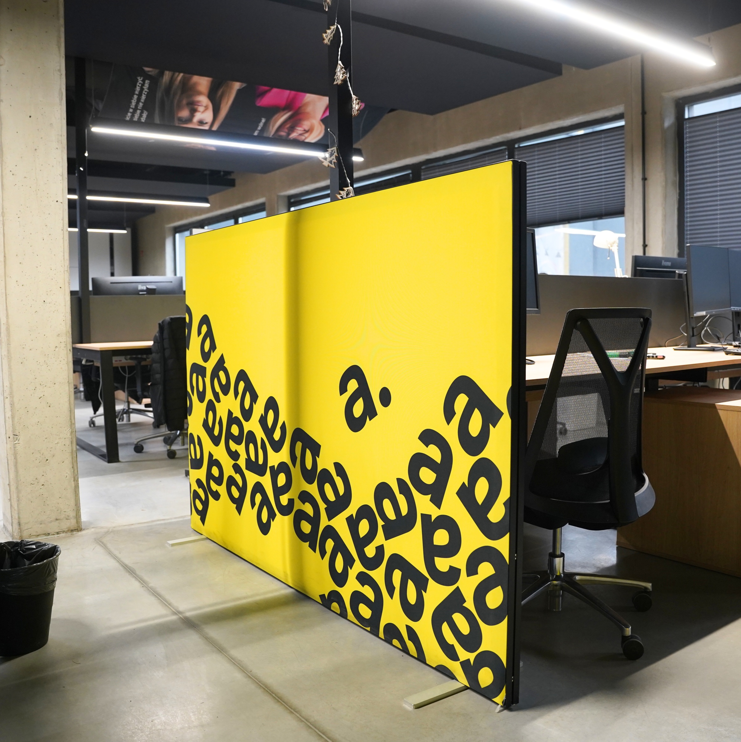 Ścianka reklamowa z żółtą grafiką i czarnymi literami „a” w biurze typu open space, ustawiona między stanowiskami pracy jako przegroda wizualna.