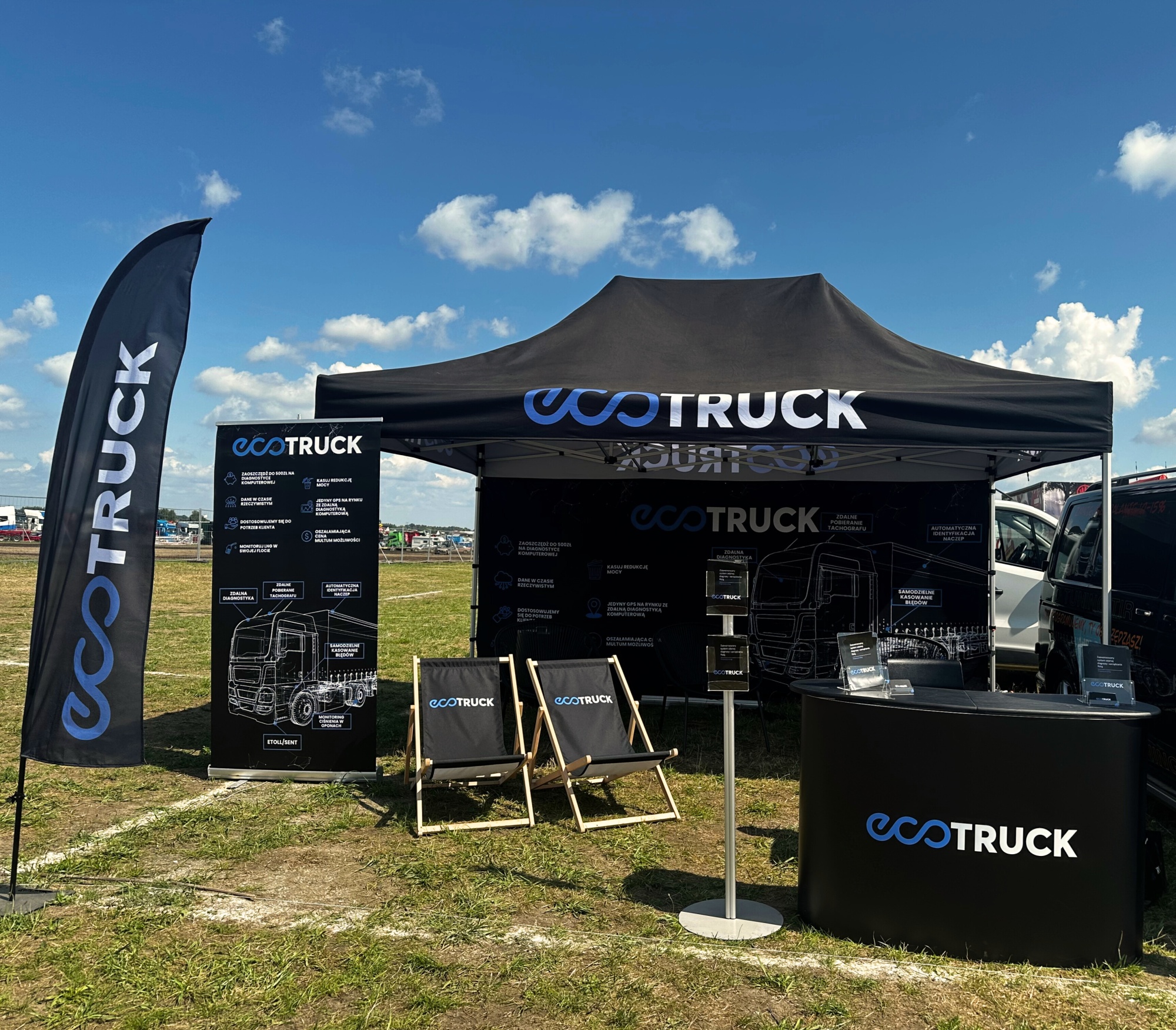 Stoisko sponsorskie EcoTruck na wydarzeniu plenerowym – namiot z nadrukiem, flagi reklamowe, trybunka, leżaki i roll-up z grafiką techniczną ciężarówki, ustawione na otwartym terenie pod niebieskim niebem. 