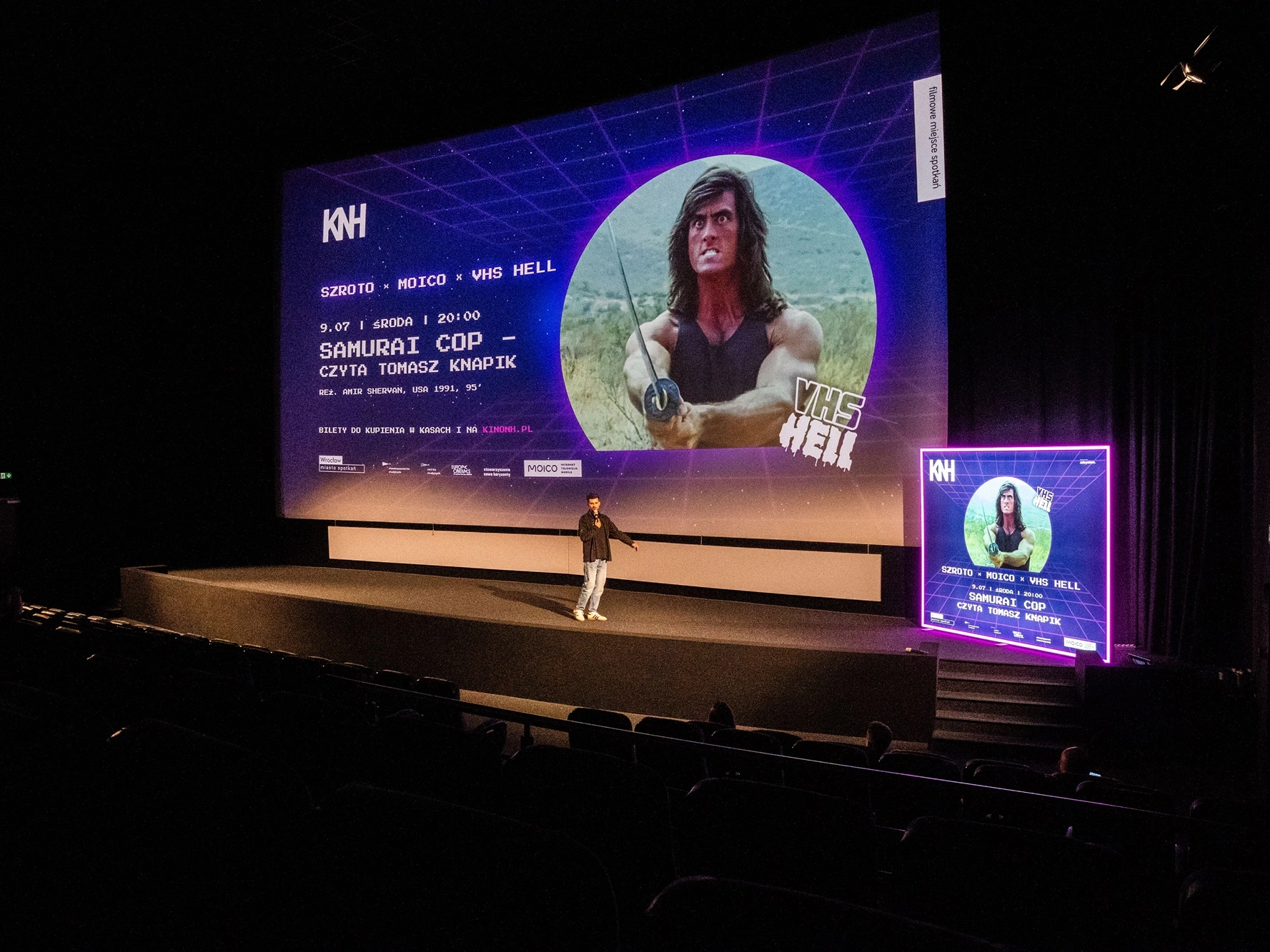 Zdjęcie przedstawia salę kinową podczas wydarzenia promującego film Samurai Cop, z dużym ekranem i podświetlanym kasetonem reklamowym ustawionym obok sceny.