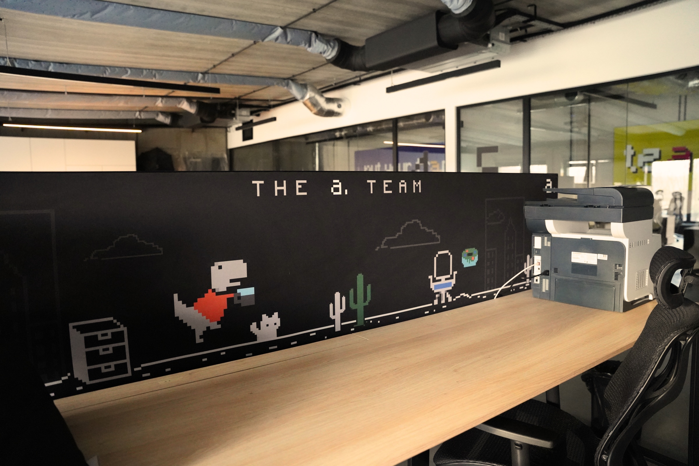 Biuro z nowoczesnym wystrojem industrialnym, stanowiskiem pracy z czarnym panelem ozdobionym grafiką w stylu pixel art z napisem "THE a. TEAM", dinozaurem i postacią z gry, obok drukarka i krzesło biurowe.