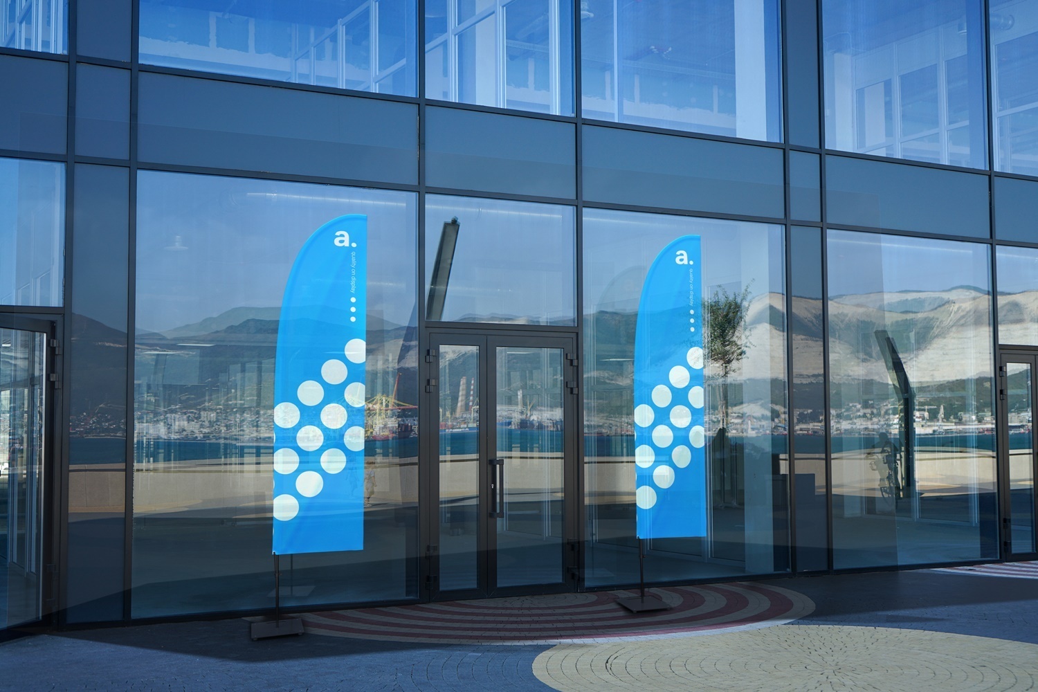 Niebieskie flagi reklamowe w kształcie łezki ustawione przed szklanym budynkiem z widokiem na góry i miasto w tle. Flagi stoją na stabilnych podstawach, tworząc elegancką ekspozycję outdoorową przed wejściem.
