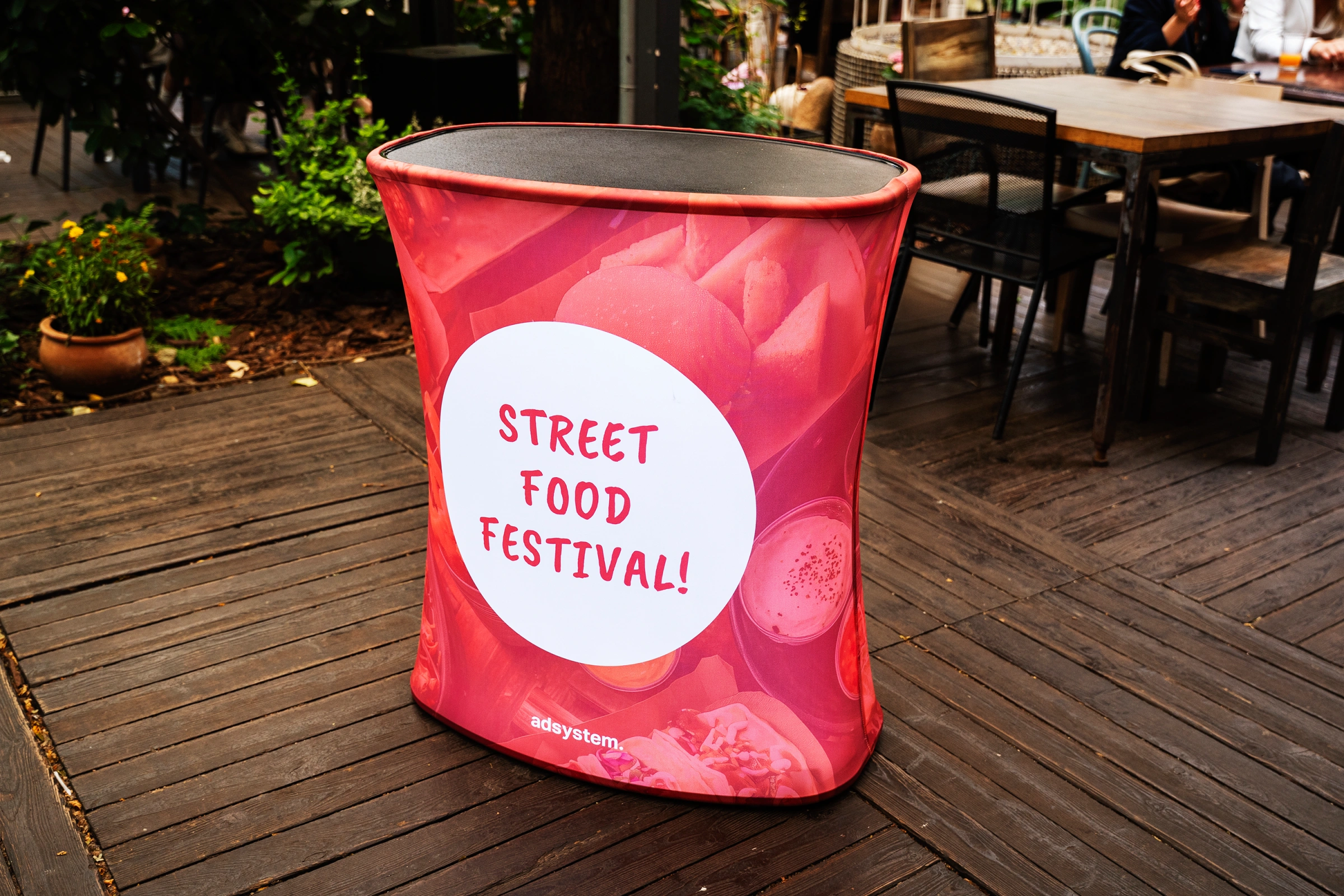 Trybunka reklamowa z nadrukiem „Street Food Festival!” ustawiona na drewnianym tarasie podczas wydarzenia plenerowego.