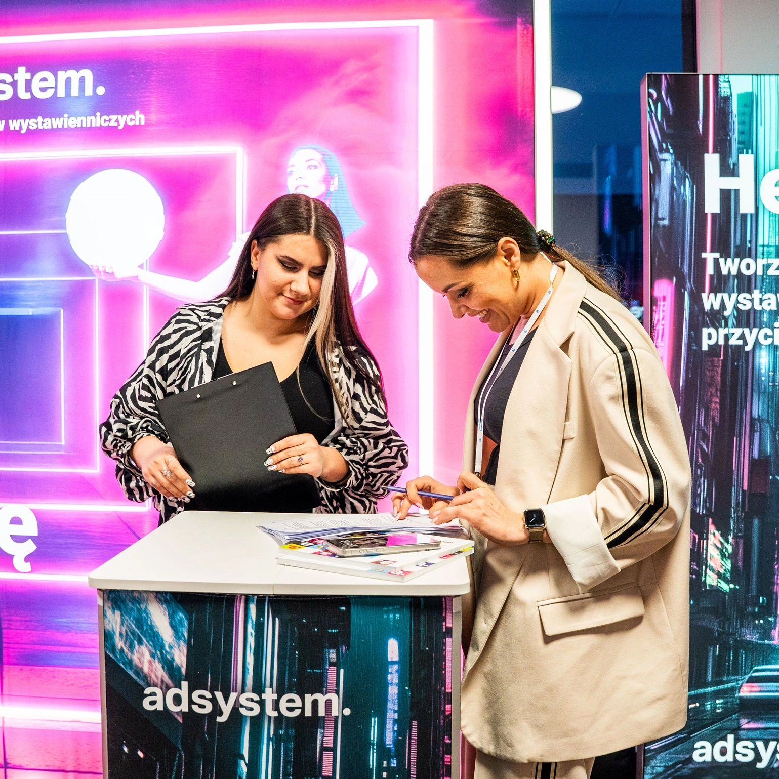 Dwie kobiety rozmawiają przy trybunce reklamowej adsystem, na tle podświetlanych systemów wystawienniczych z neonowymi grafikami i hasłami.