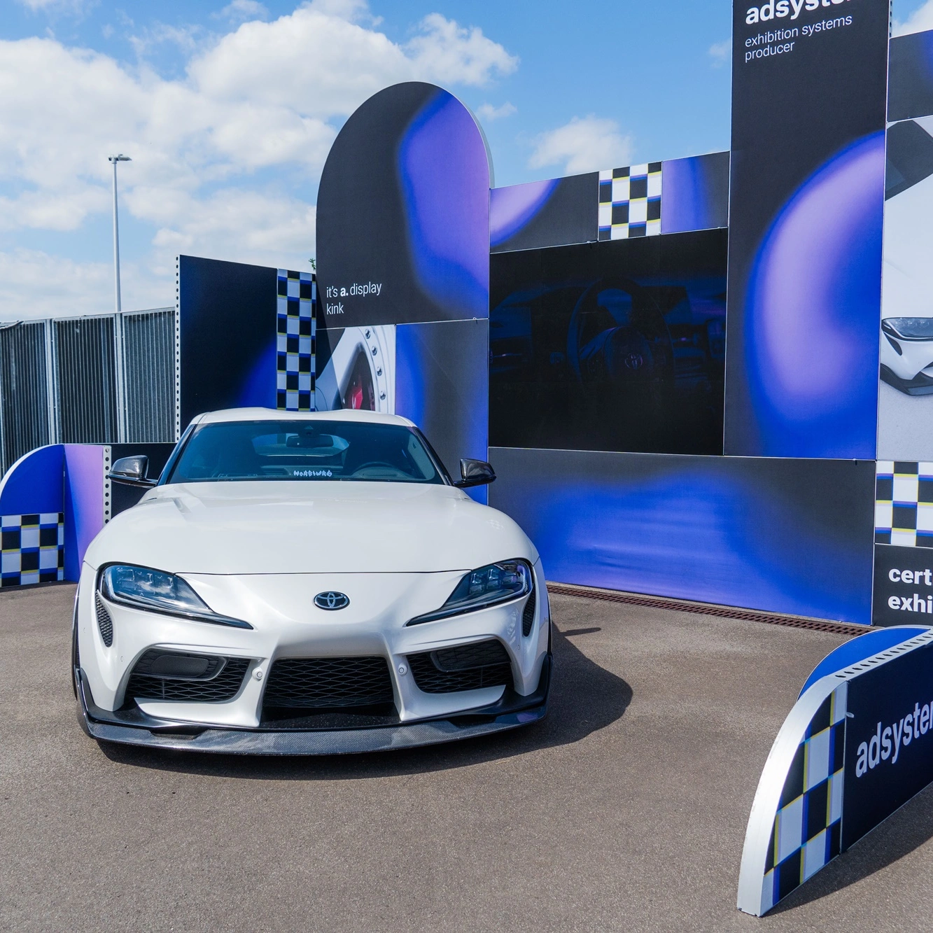 Biała Toyota Supra wyeksponowana na stoisku adsystem z czarno-fioletowymi panelami i motywem szachownicy podczas wydarzenia Ultrace 2025.