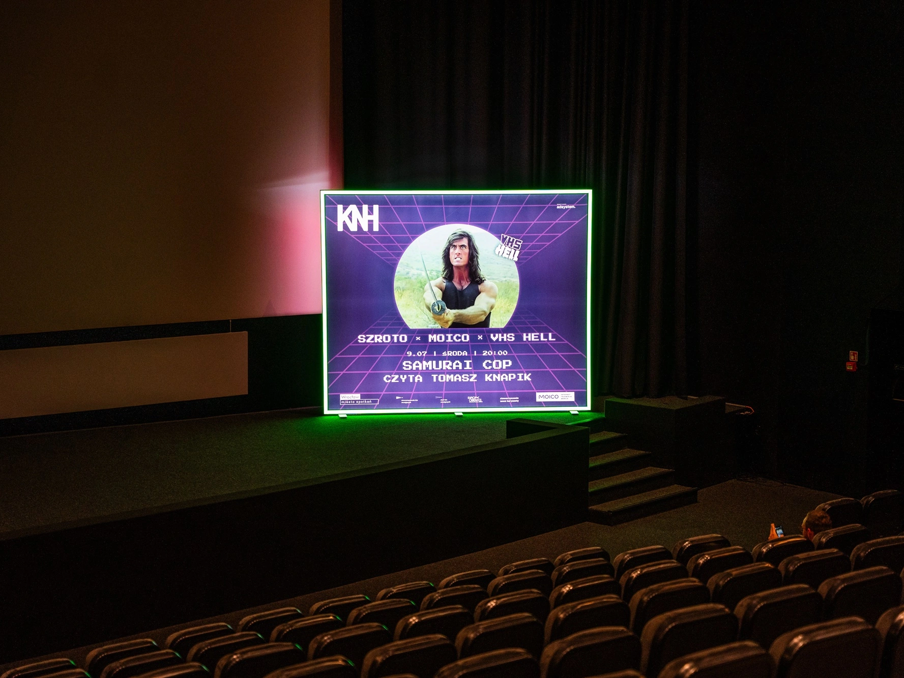 Zdjęcie przedstawia podświetlaną ściankę reklamową z grafiką promującą wydarzenie filmowe, ustawioną w sali kinowej Kina Nowe Horyzonty.
