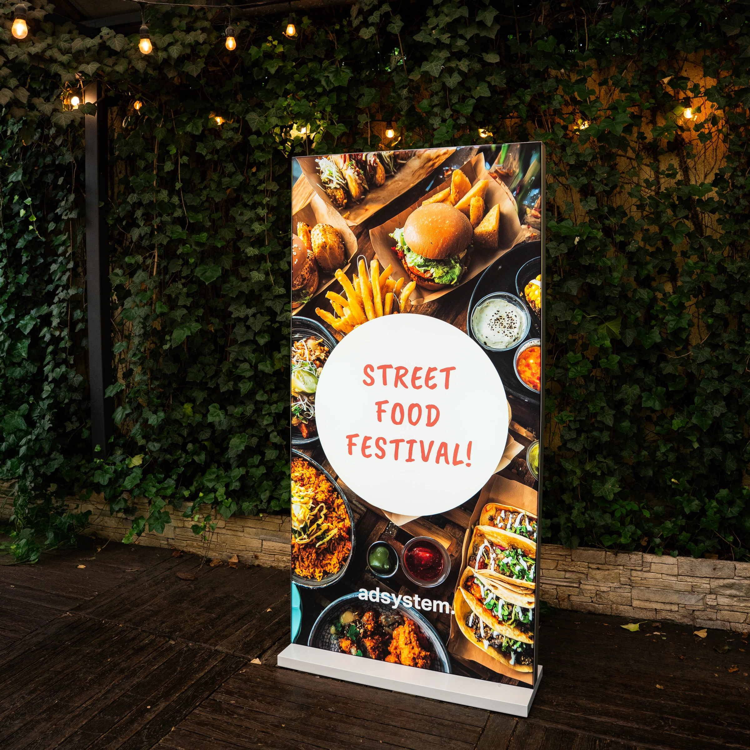 Ścianka reklamowa z nadrukiem „Street Food Festival!” przedstawiająca kolorowe dania, ustawiona na tle zielonej ściany z bluszczu i lampek dekoracyjnych.