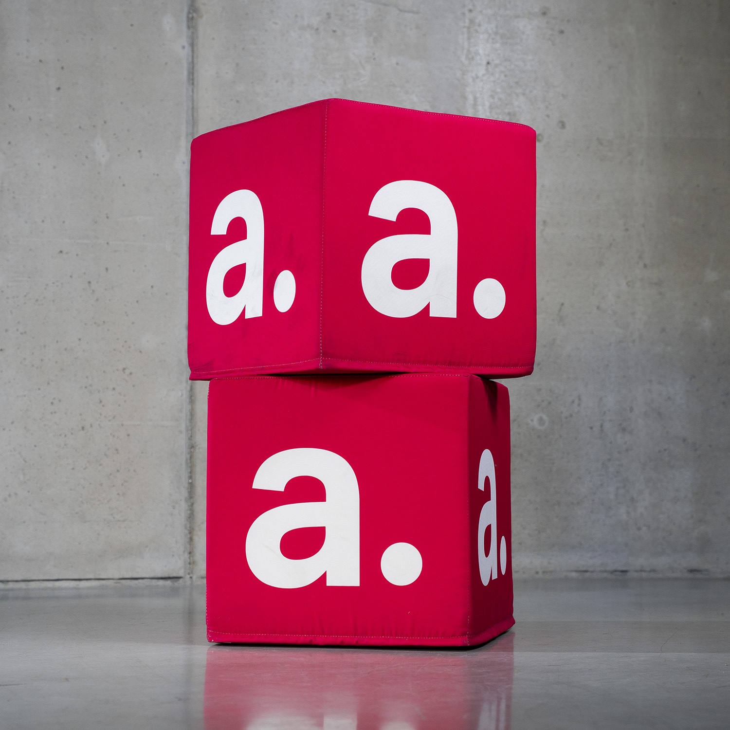 Dwie pufy kostki z wymiennymi, czerwonymi pokrowcami z nadrukiem „a.”, gotowe do ponownego wykorzystania w różnych aranżacjach.