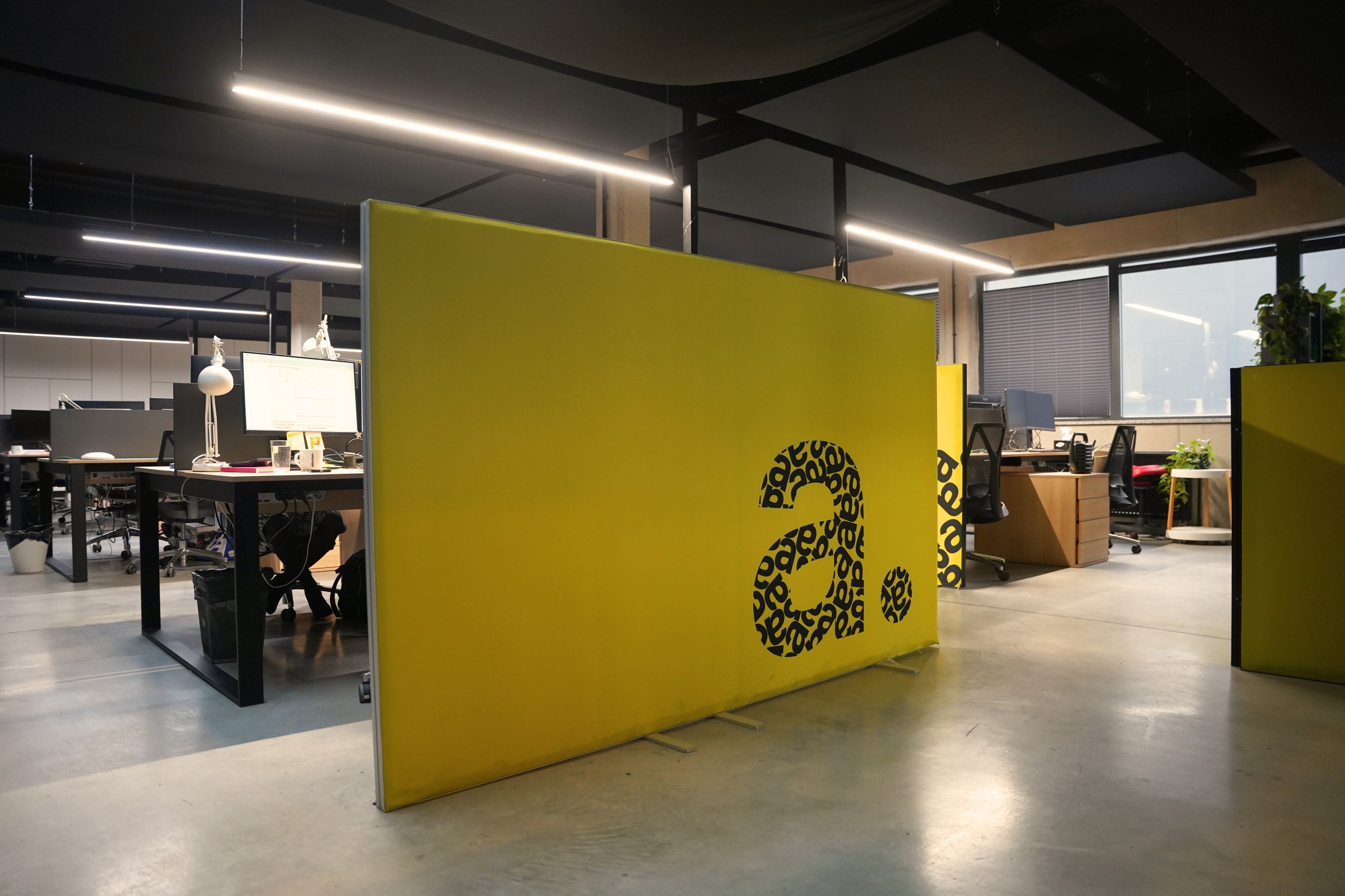 Przestrzeń biurowa z nowoczesnymi stanowiskami pracy, oddzielona żółtą ścianką z logotypem „a.” w graficznym wzorze; widoczne biurka, monitory i oświetlenie sufitowe LED.