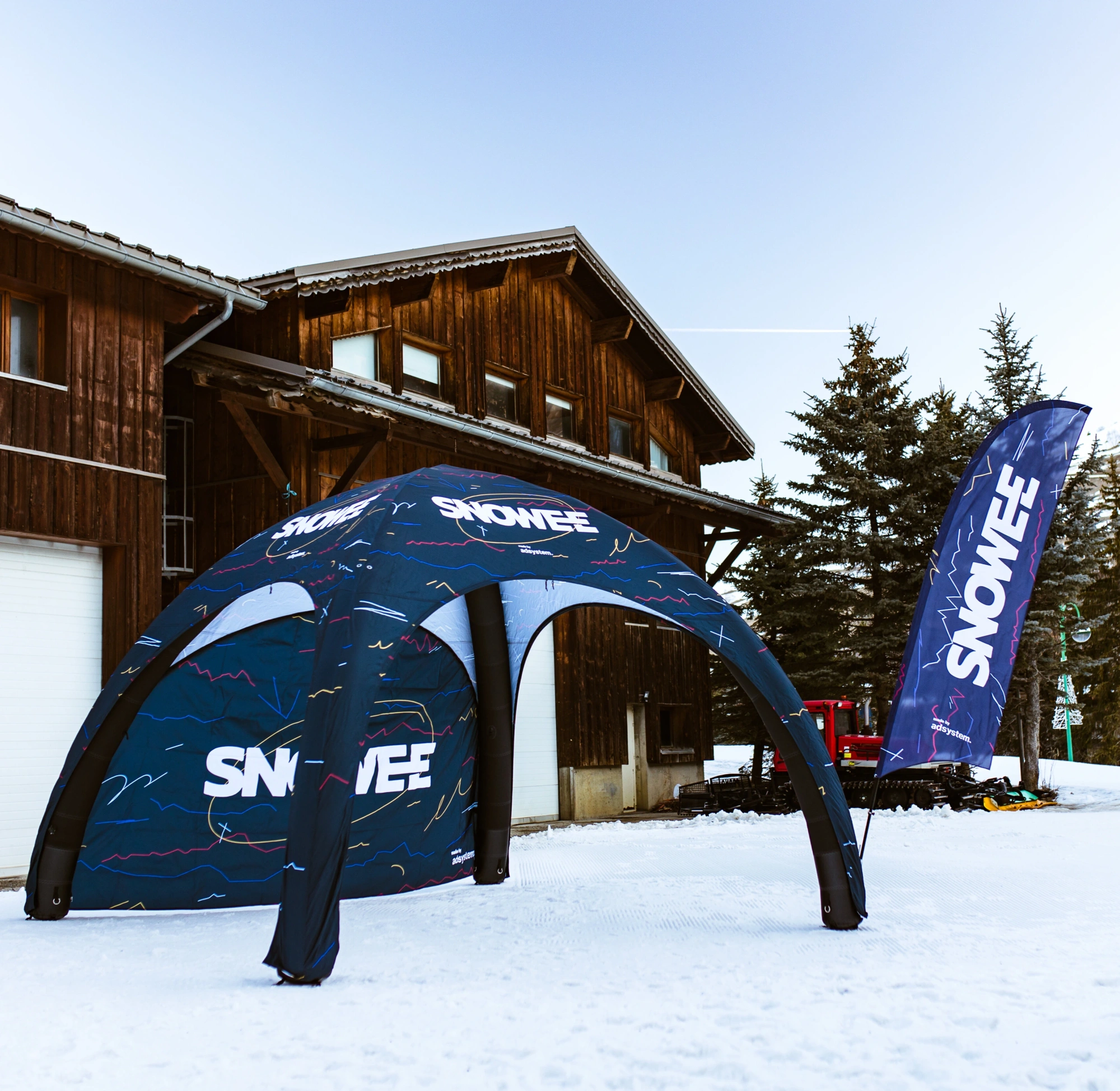 Namiot stałociśnieniowy i flaga reklamowa z logo Snowee podczas zimowego wydarzenia sportowego w Alpach, z widocznym ośnieżonym terenem i drewnianym budynkiem w tle.