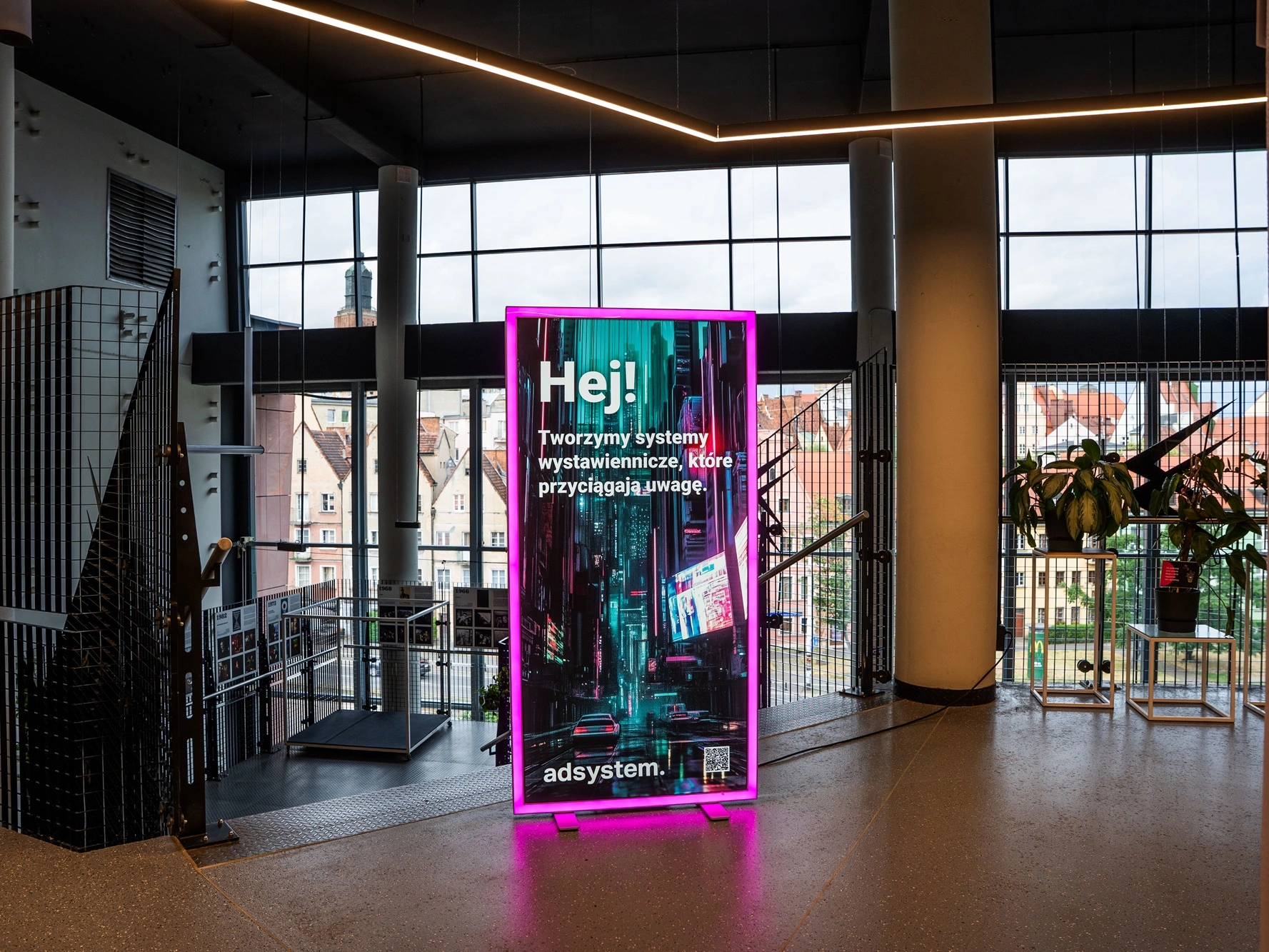 Zdjęcie przedstawia podświetlaną ściankę reklamową adsystem ustawioną w przestrzeni z dużymi oknami, przez które widać panoramę miasta.