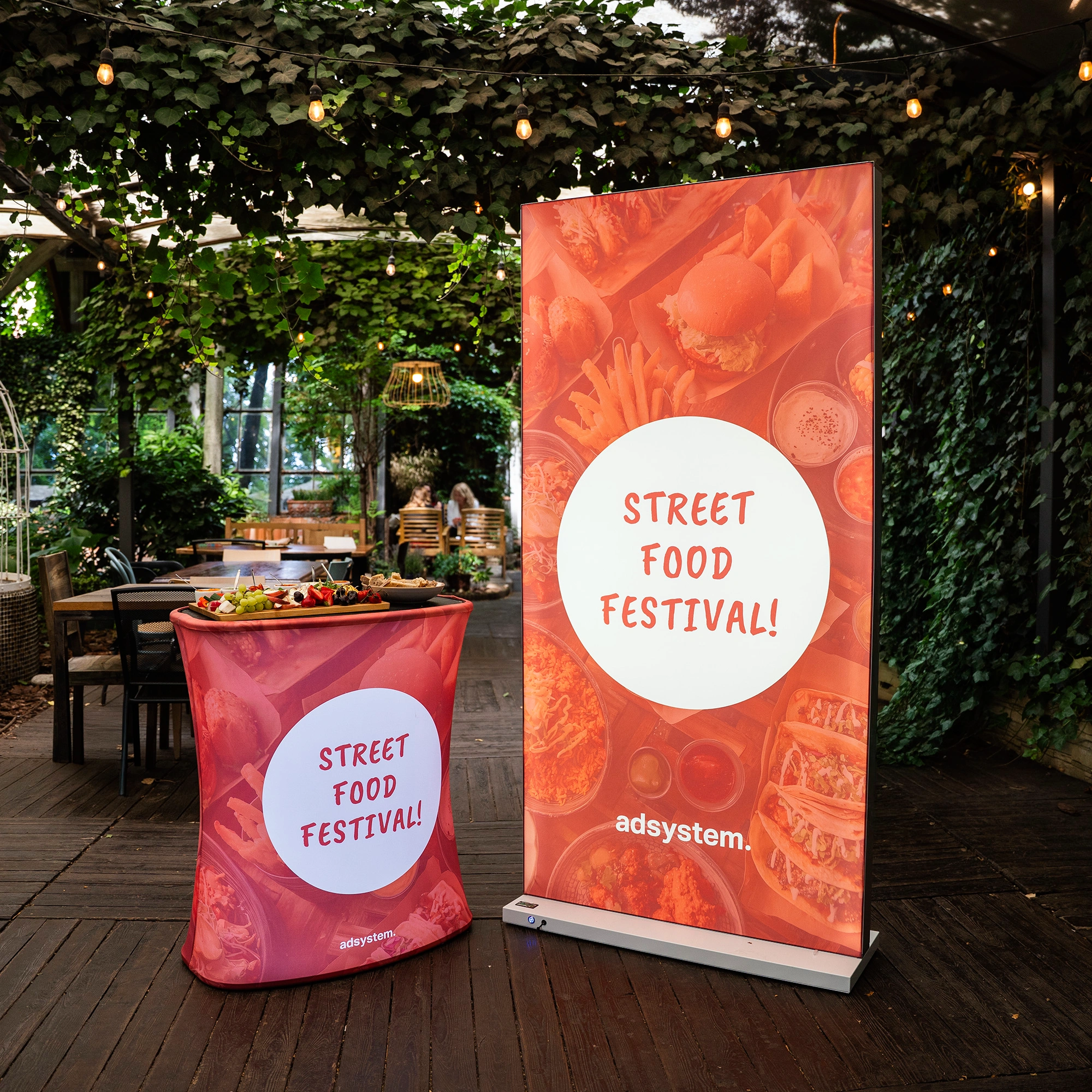 Trybunka i ścianka reklamowa z motywem „Street Food Festival!” ustawione w ogrodowej przestrzeni eventowej pod dekoracyjnymi lampkami.